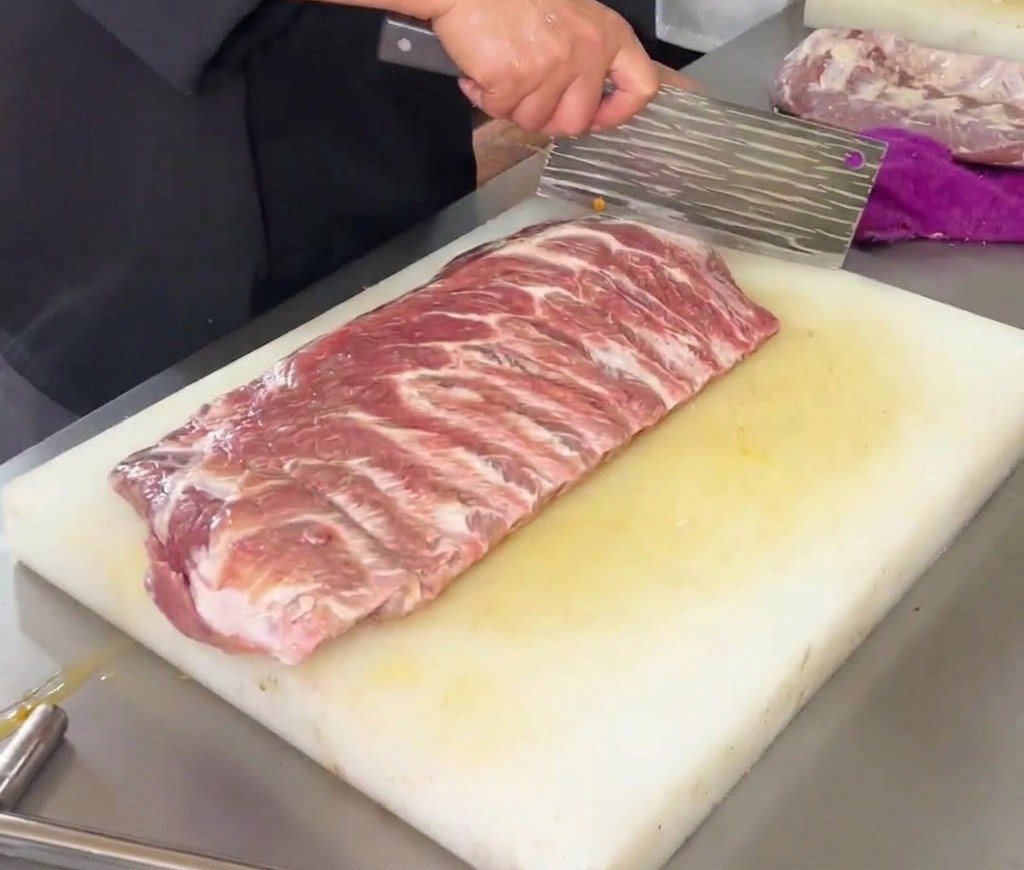 Uno chef che usa una grande mannaia da cucina per incidere un motivo a rombi su un pezzo di costine di maiale crude su un tagliere di plastica bianca.