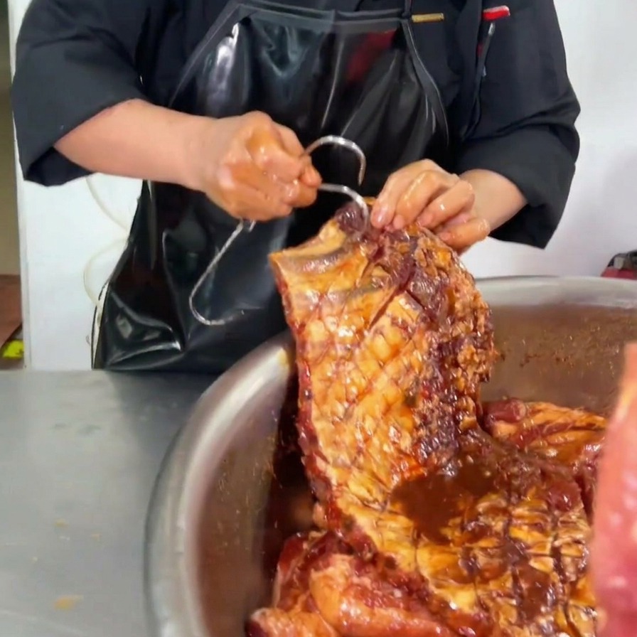 Uno chef che inserisce un grande gancio metallico nellangolo superiore di un pezzo di costine di maiale completamente marinato e coperto di salsa scura sopra una ciotola di acciaio.