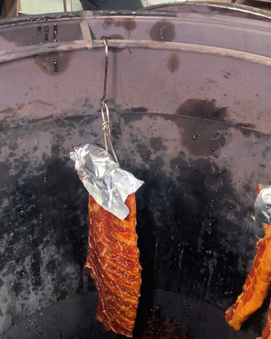 Un pezzo di costine di maiale marinate con un foglio di alluminio a protezione della parte superiore appeso allinterno di un forno cilindrico profondo e scuro.