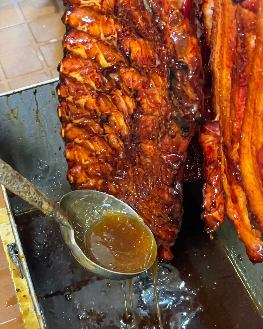 Un pezzo di costine di maiale lucido e glassato al miele appeso verticalmente sopra una vaschetta metallica mentre la glassa dolce in eccesso cola verso il basso.