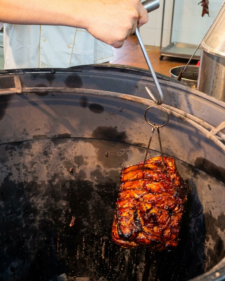 Un pezzo di costine di maiale lucido che viene inserito tramite un gancio metallico in un forno cilindrico profondo e scuro.