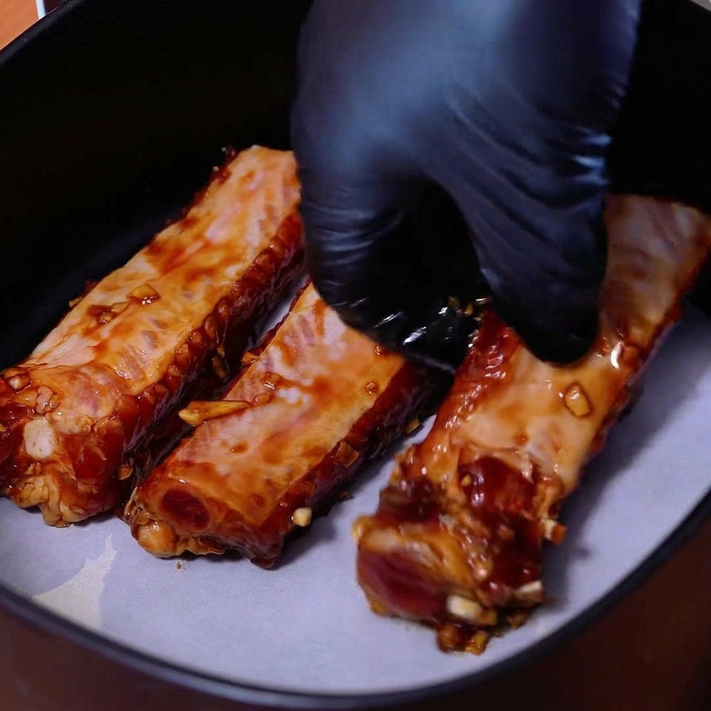 Costine di maiale marinate disposte ordinatamente una accanto allaltra su carta forno nel cestello della friggitrice ad aria.