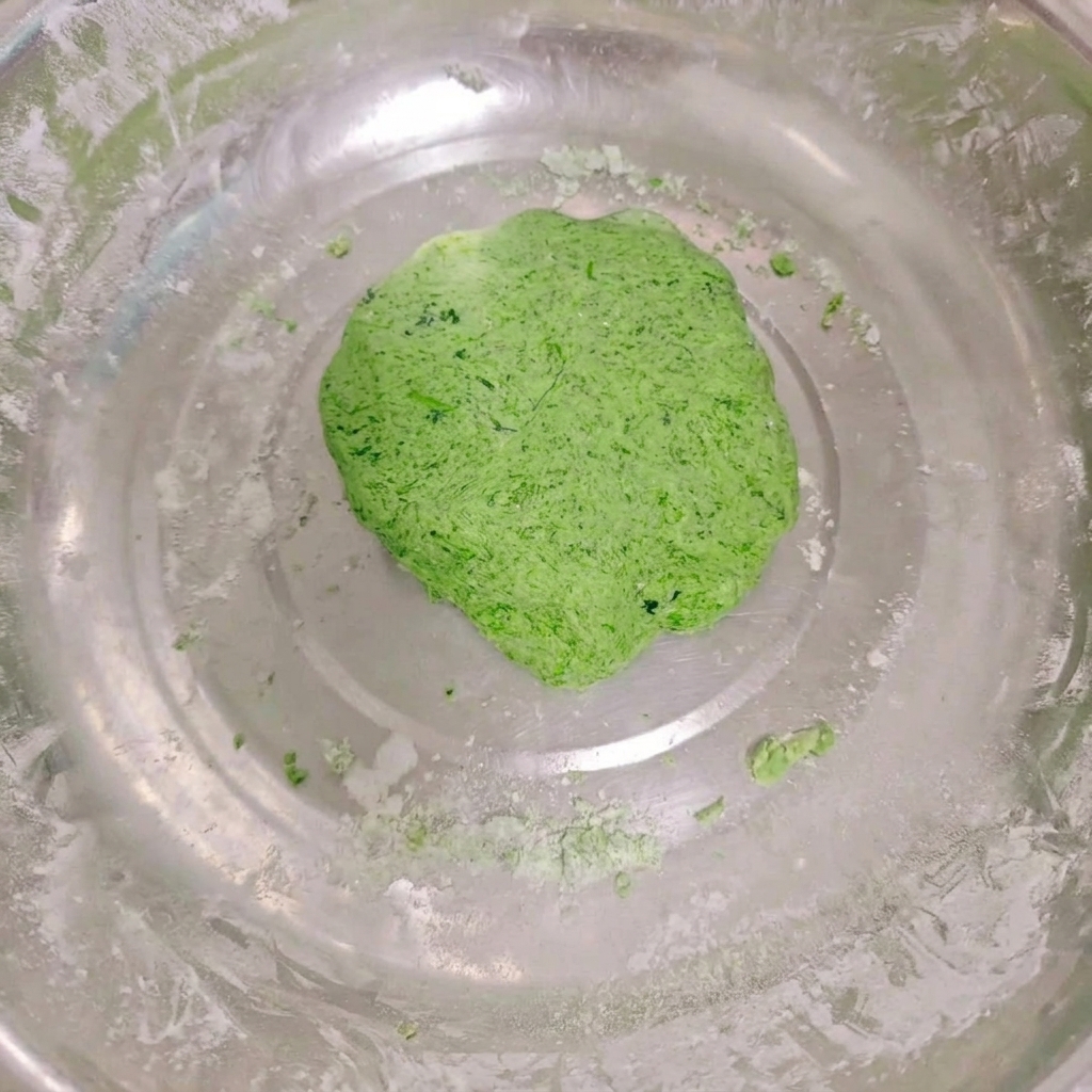 Un panetto di verdure liscio e dal colore verde acceso allinterno di una ciotola capiente in acciaio inossidabile, con tracce di farina sui bordi.
