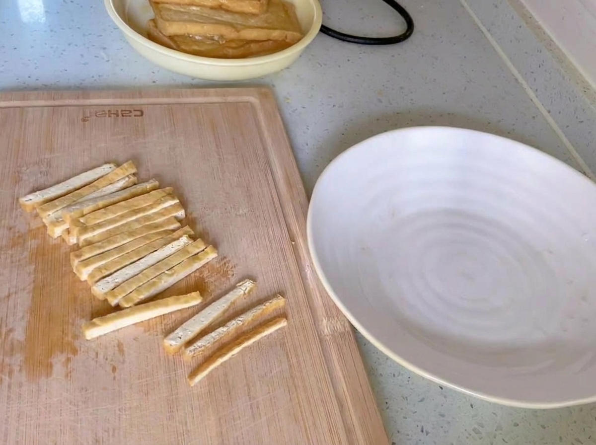 Tofu fritto dorato tagliato a strisce sottili e uniformi su un tagliere di legno accanto a un piatto bianco.