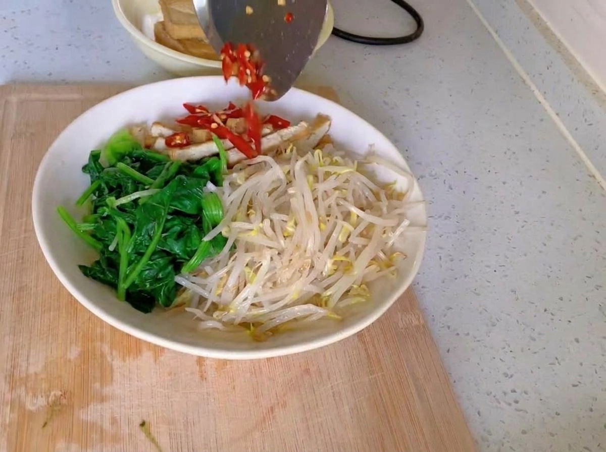 Una ciotola bianca riempita con sezioni distinte di spinaci verdi, germogli di soia chiari e strisce di tofu, sormontati da un mucchietto di peperoncini rossi tritati e aglio sminuzzato.