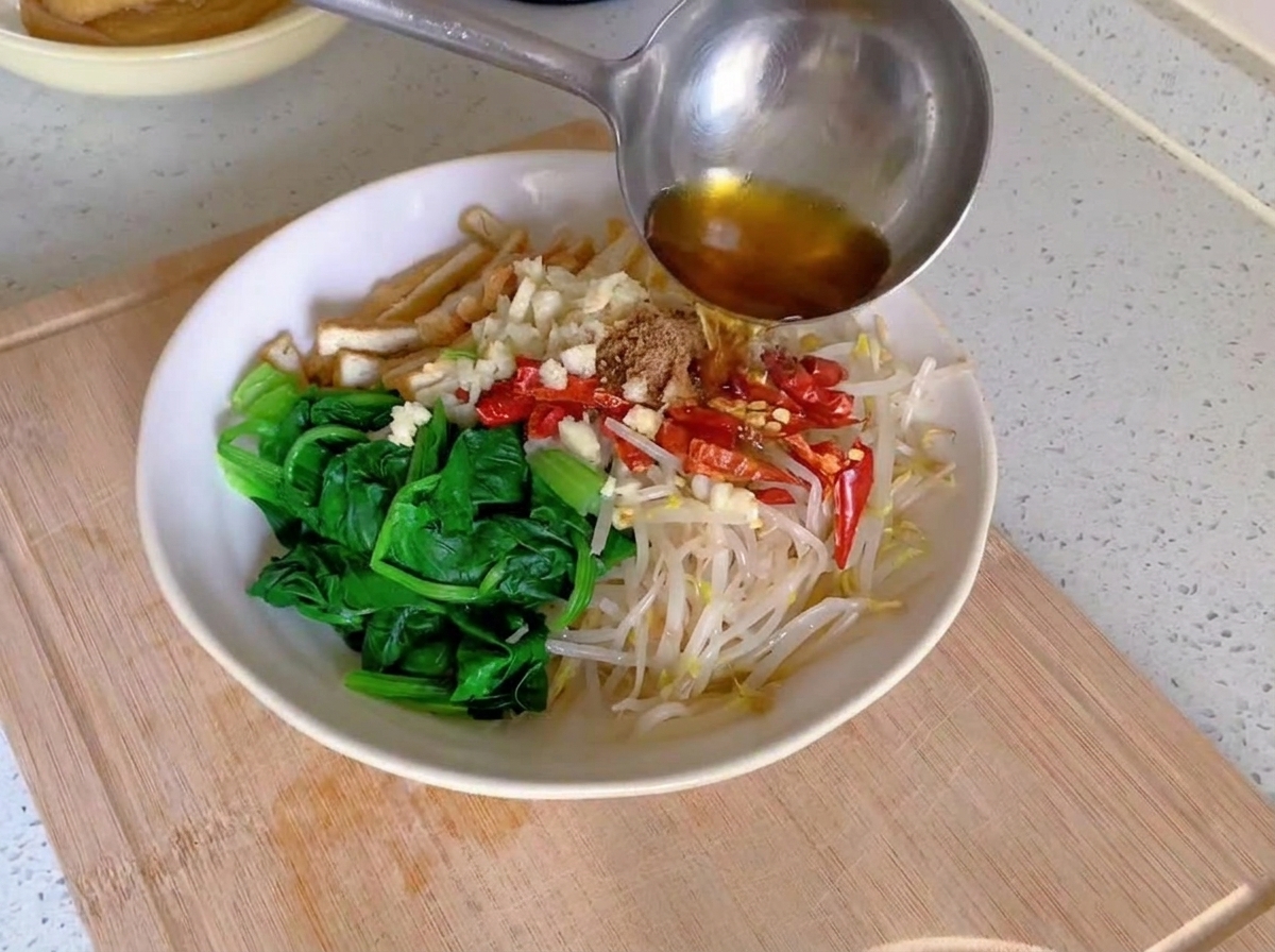 Olio bollente e fumante versato da un mestolo di metallo su aglio tritato e peperoncini rossi secchi disposti sopra le verdure sbollentate in una ciotola bianca.