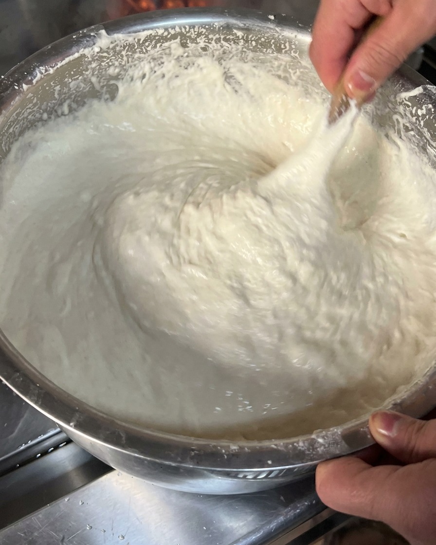 Un primo piano di un impasto bianco, denso e appiccicoso, sollevato e teso con un utensile di legno allinterno di una ciotola di metallo.