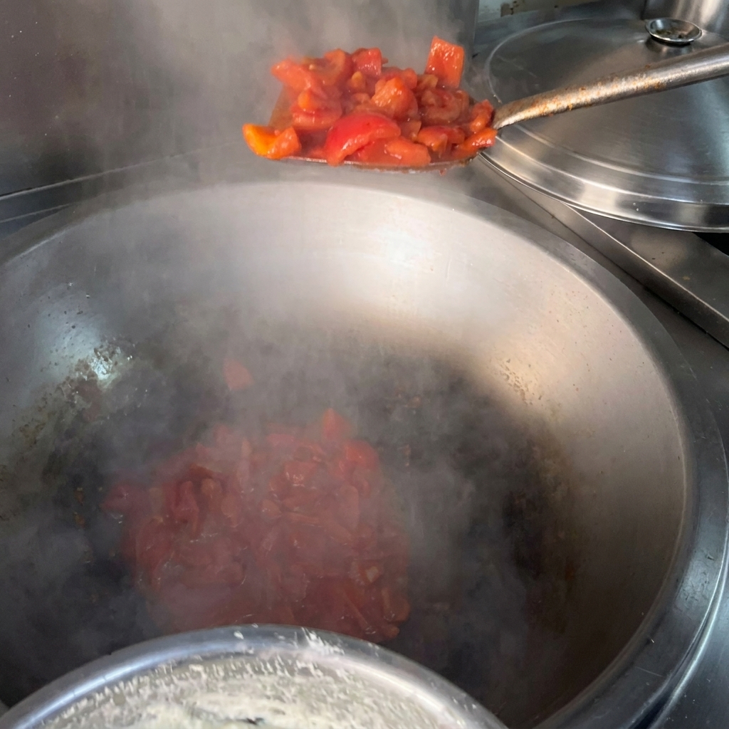 Una spatola di metallo che aggiunge pomodori rossi a pezzetti in un grande wok fumante con olio caldo.