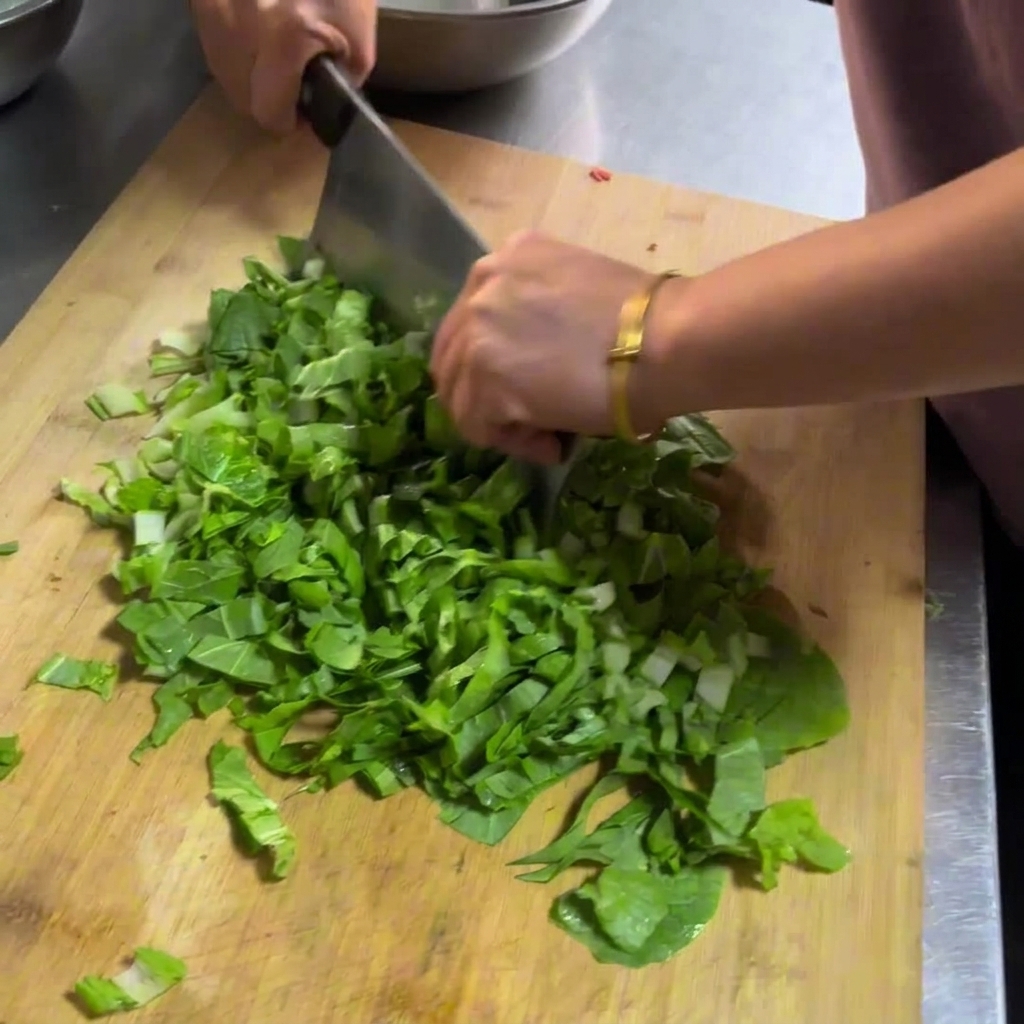 Mani che usano una grande mannaia di metallo per tagliare verdure fresche a foglia verde su un tagliere di legno.