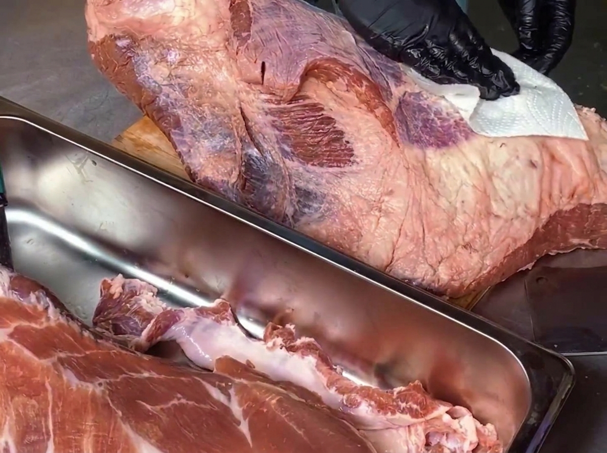 Un cuoco con guanti neri usa della carta da cucina per tamponare un grande pezzo di punta di petto di manzo cruda su un tagliere di legno.