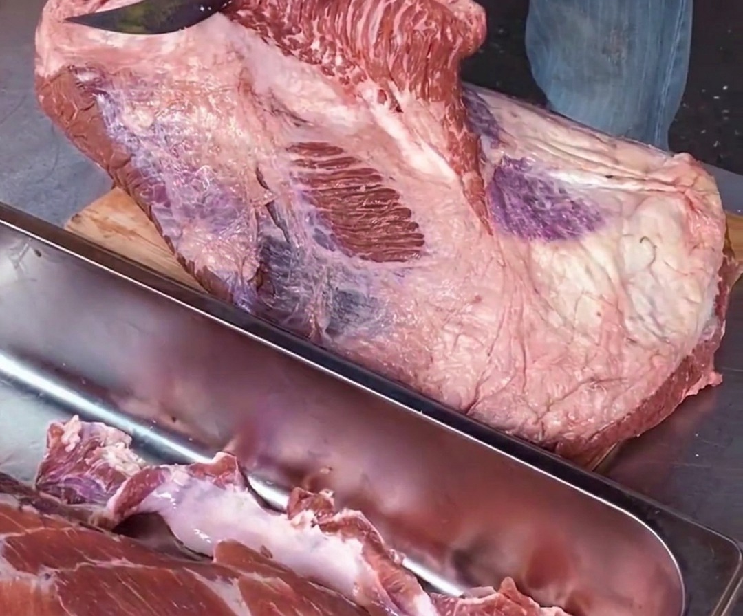 Primo piano di uno chef che usa un coltello affilato parallelamente al tagliere per separare la parte del point da quella del flat lungo una venatura di grasso bianco.