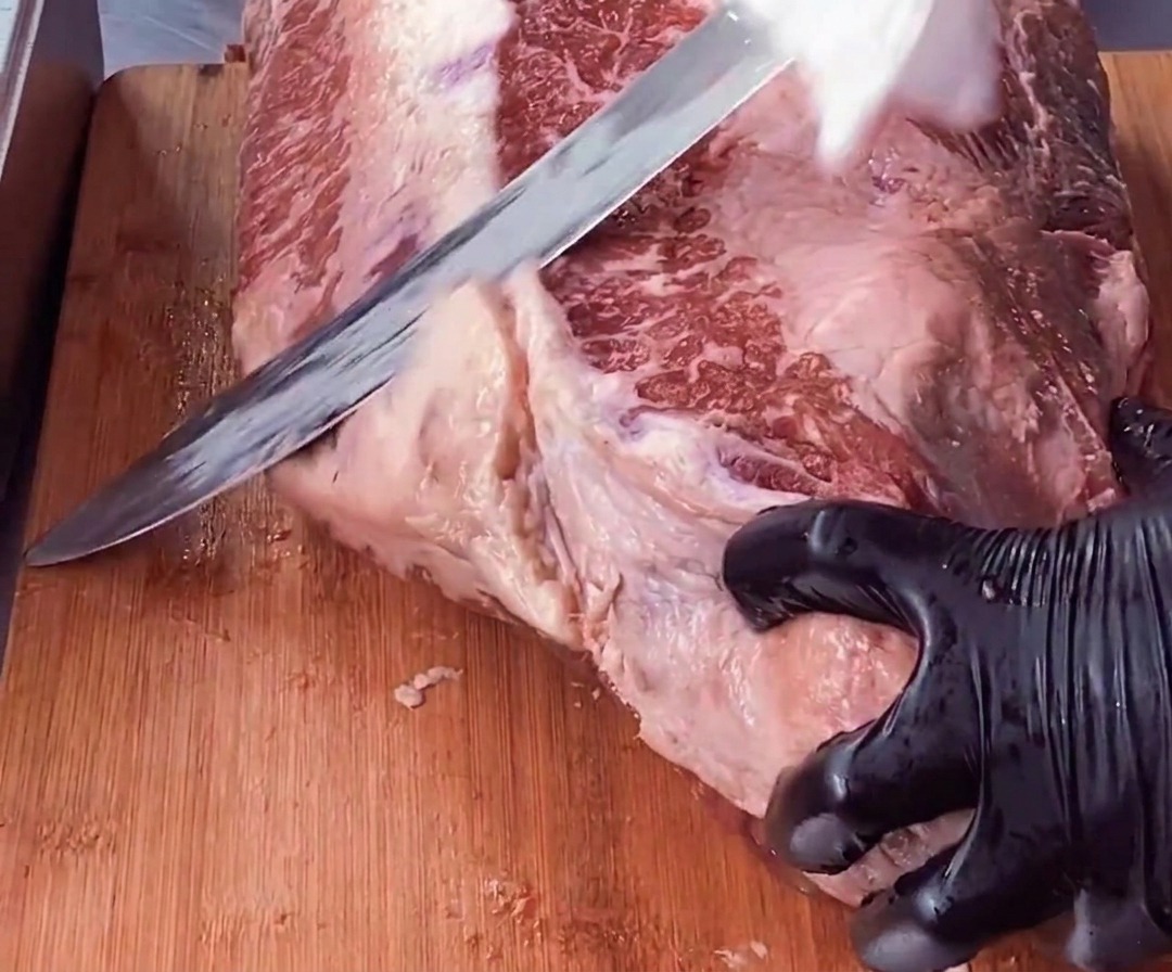 Una mano protetta da un guanto tiene fermo un pezzo di brisket mentre un lungo coltello rimuove uno spesso strato di grasso bianco dalla carne rossa.