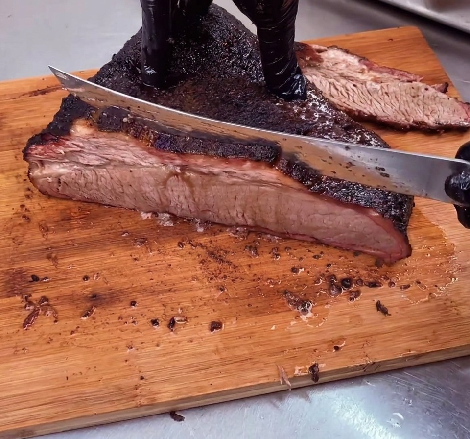 Uno chef affetta un brisket perfettamente affumicato con una crosta scura, tagliandolo perpendicolarmente alle fibre su un tagliere di legno.