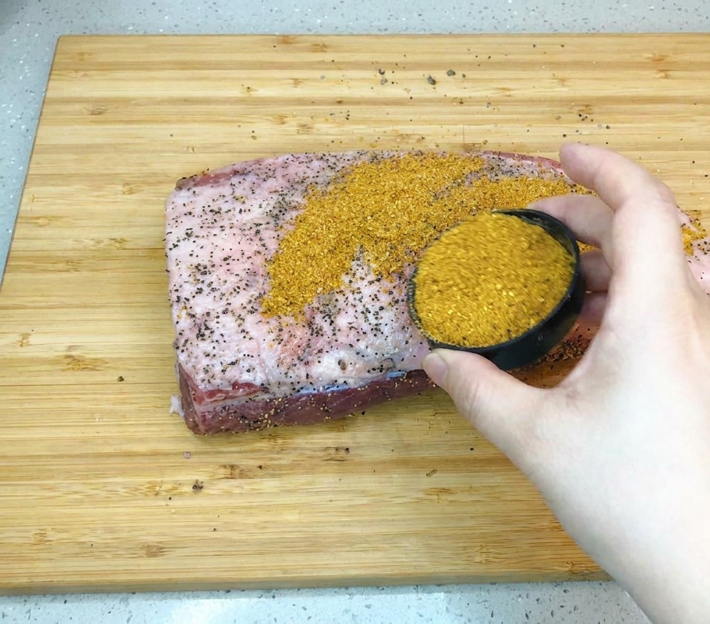 Una mano versa un mix di spezie per barbecue color ambra su un grande pezzo di punta di petto di manzo crudo appoggiato su un tagliere di legno.