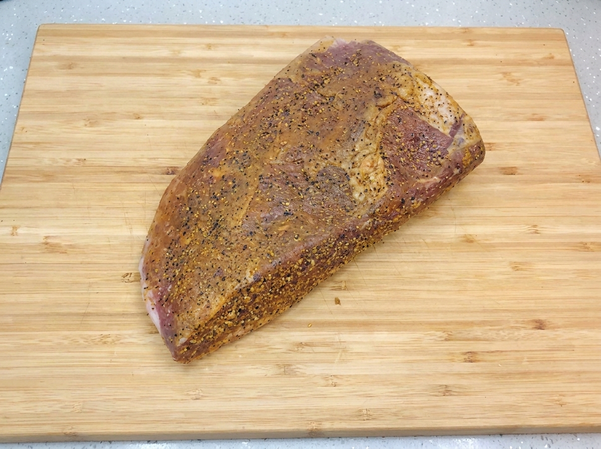 Il brisket marinato e speziato riposa su un tagliere di legno chiaro, lucido per un velo dolio e cosparso di pepe nero macinato fresco.