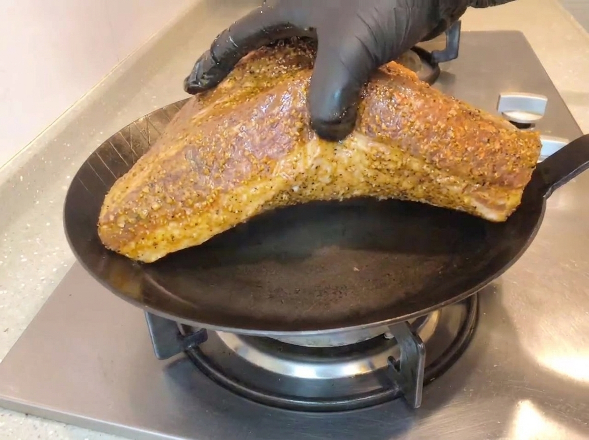 Una mano con un guanto nero adagia il grande pezzo di manzo dal lato del grasso in una padella di ghisa ben calda su un fornello a gas.