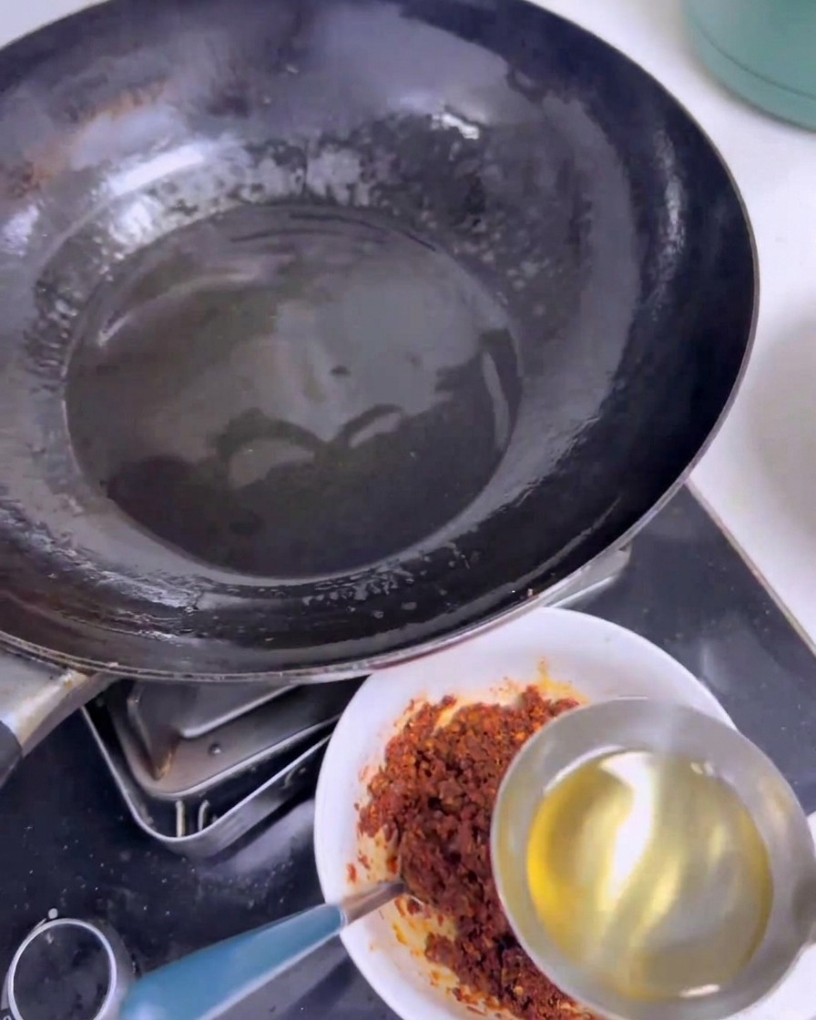 Un mestolo di acciaio versa olio bollente e limpido in una ciotola bianca contenente spezie e peperoncino rosso, accanto a un wok nero.