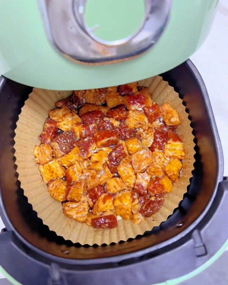 Pezzetti marinati di grasso di manzo disposti in un unico strato su carta da forno allinterno del cestello verde di una air fryer.