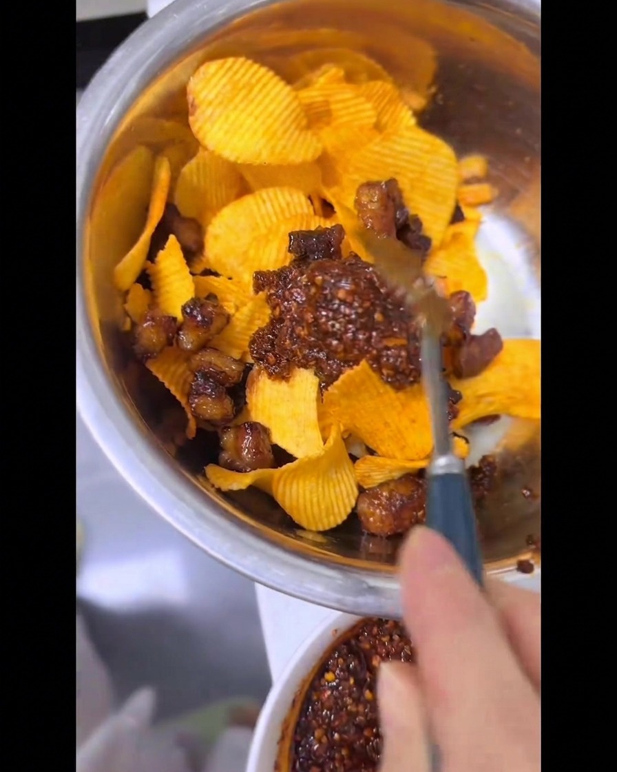 Una mano usa un cucchiaio per amalgamare patatine rigate, grasso di manzo croccante e un filo di olio rosso piccante in una bacinella di metallo.