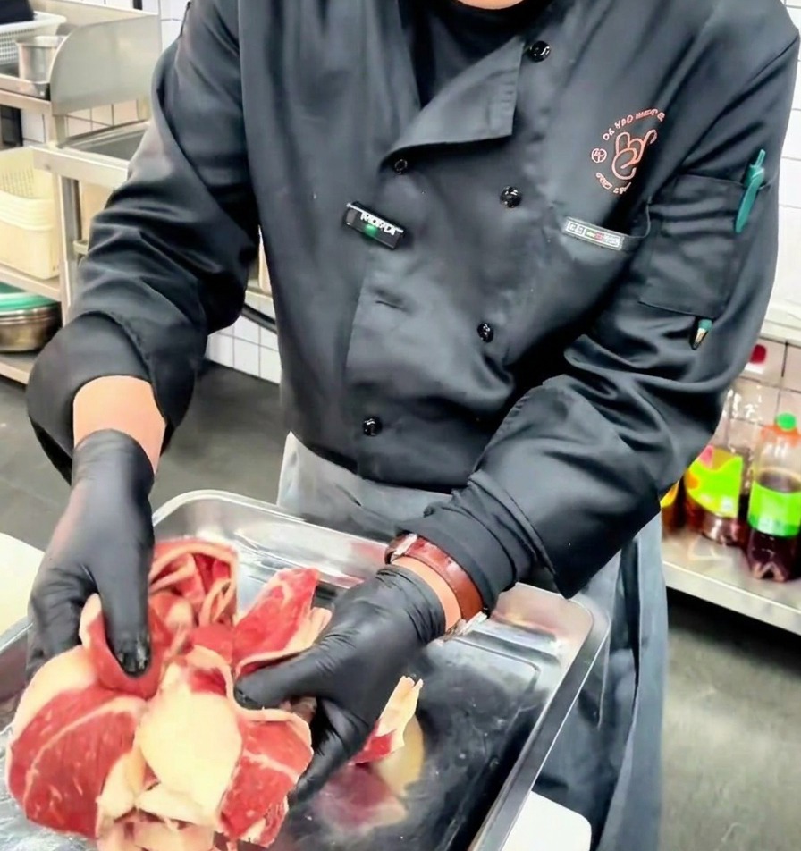 Uno chef con guanti neri mostra sottili fette di punta di petto di manzo cruda con strati ben definiti di grasso e polpa su un vassoio di metallo.