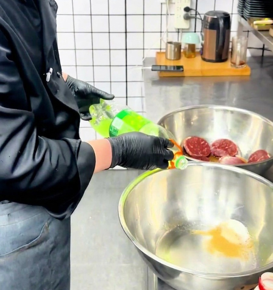 Uno chef versa un liquido verde chiaro da una bottiglia in una ciotola di acciaio inossidabile contenente le spezie in polvere.