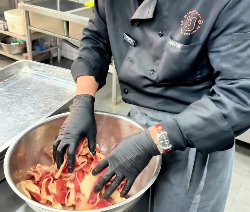 Uno chef mescola e massaggia con le mani in guanti neri le fette sottili di manzo crudo con la marinatura in una grande ciotola di acciaio.