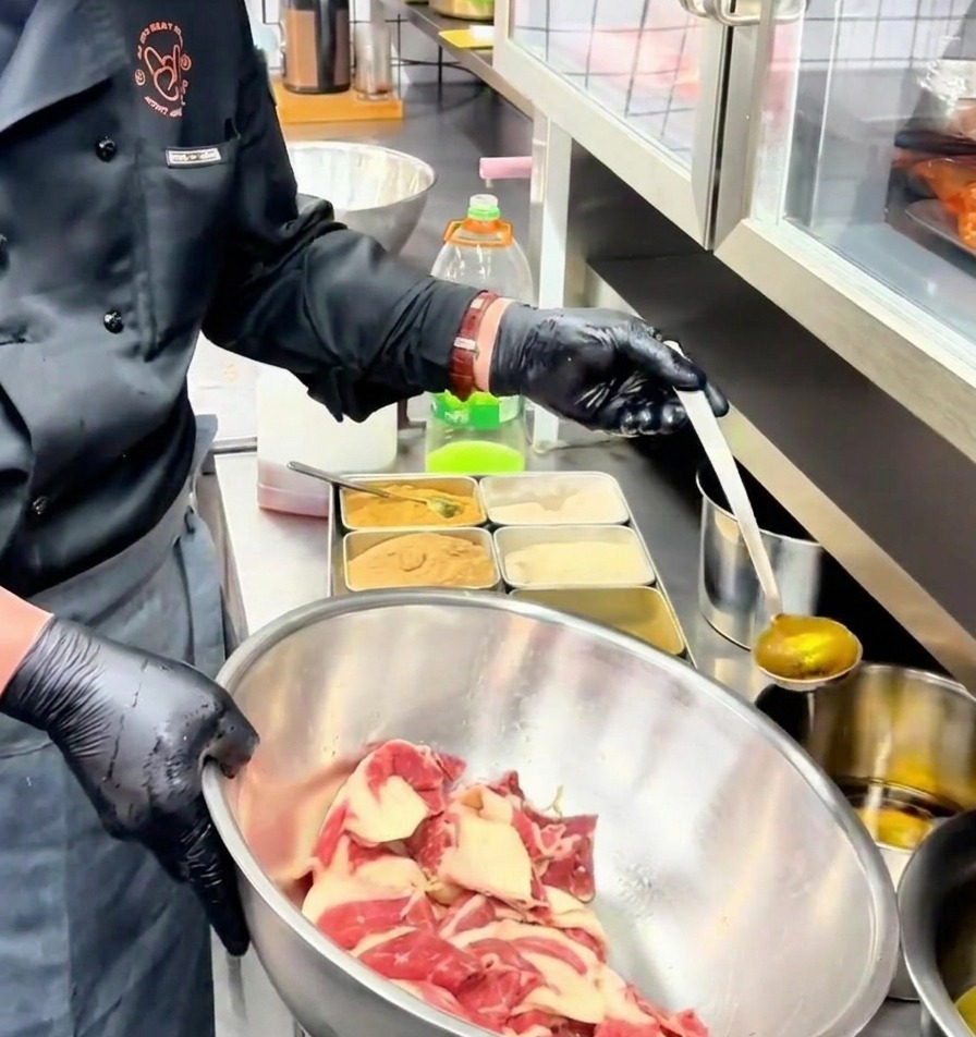 Un mestolo versa dellolio aromatizzato dorato sulle fette di manzo marinate allinterno di una ciotola di acciaio inossidabile.