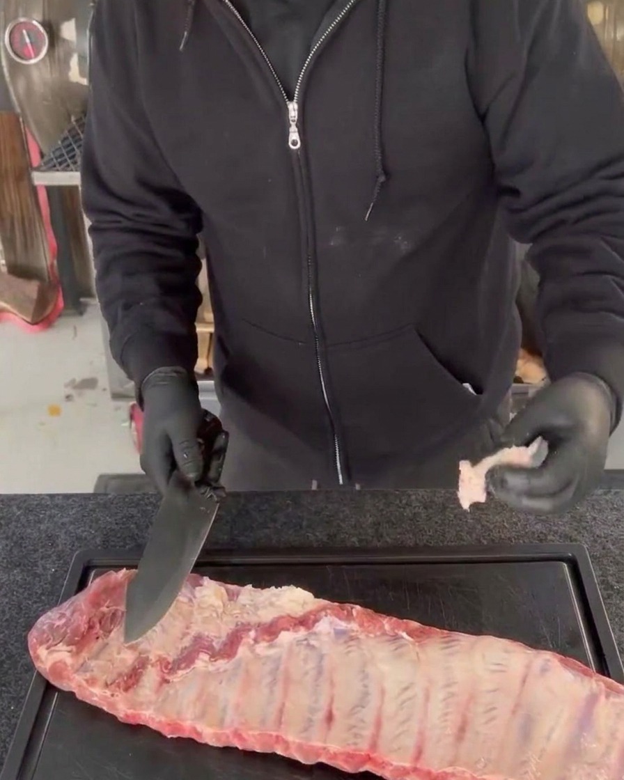 Una persona con guanti neri refila la carne in eccesso e i bordi irregolari da un baffo crudo di costine di maiale usando un grande coltello da chef su un tagliere nero.