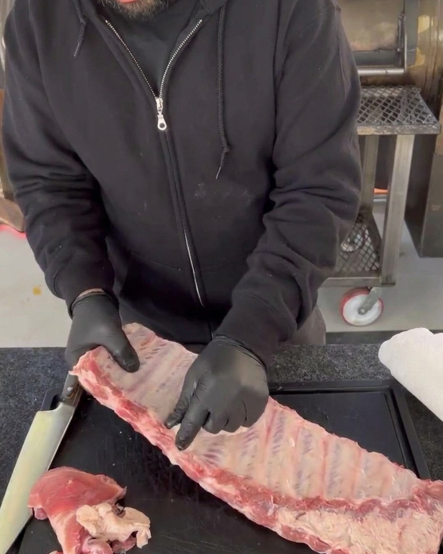 Mani con guanti che rimuovono la membrana lucida e trasparente dal retro delle ossa di un baffo crudo di costine di maiale su un tagliere.