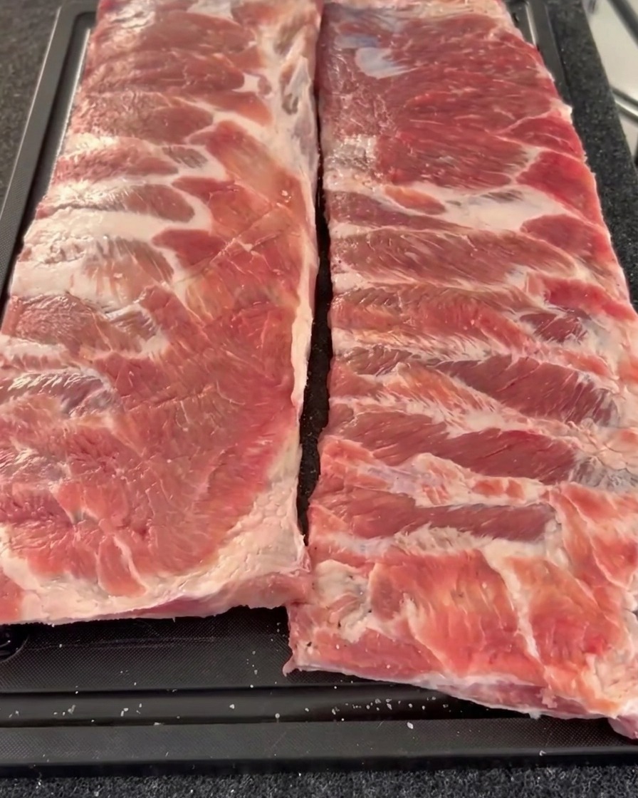 Due baffi crudi di costine di maiale disposti uno accanto allaltro su un tagliere, che mostrano il lato polposo condito con pepe grosso e sale.