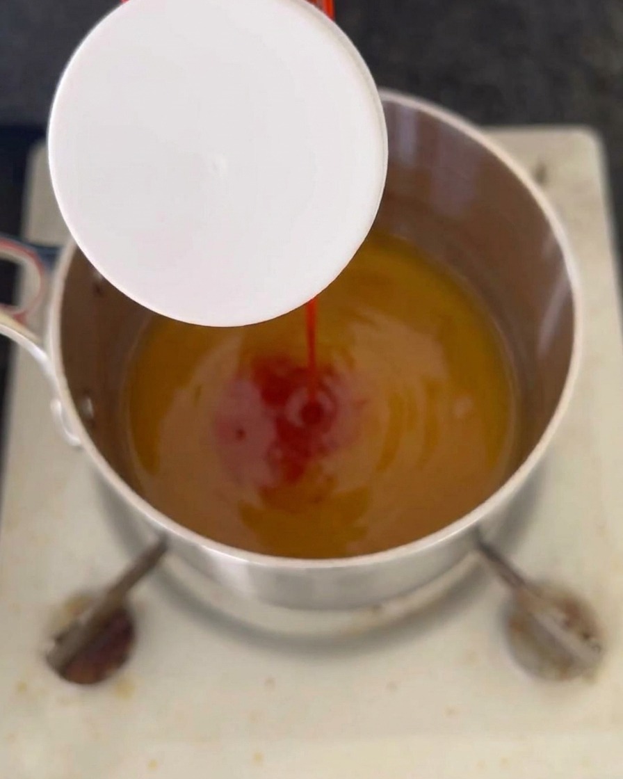 Un flusso di ketchup rosso versato in un liquido ambrato che sobbolle allinterno di un pentolino in acciaio inossidabile sui fornelli.