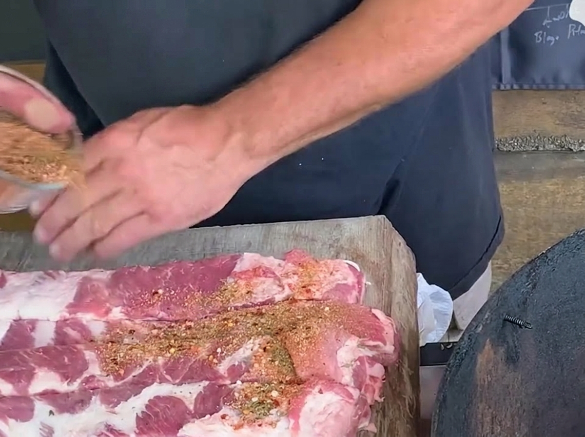 Una persona cosparge una miscela di spezie da una ciotola di vetro su lunghe strisce di costine di maiale crude su un tagliere di legno rustico.