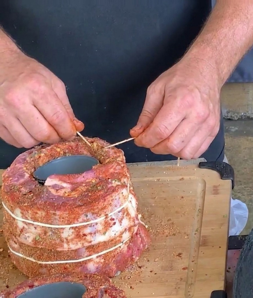 Mani che usano spago da cucina bianco e uno stuzzicandenti per legare e fissare un grande rotolo cilindrico di costine di maiale condite avvolte intorno a una lattina.