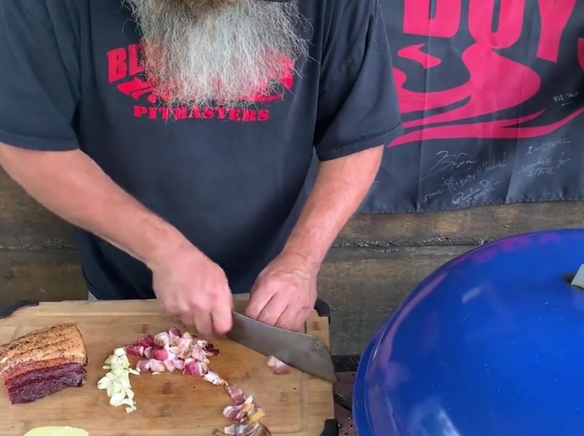 Un uomo con una lunga barba grigia usa una grande mannaia da cucina per tritare pezzi di bacon e spicchi daglio su un tagliere di legno accanto a un barbecue blu.