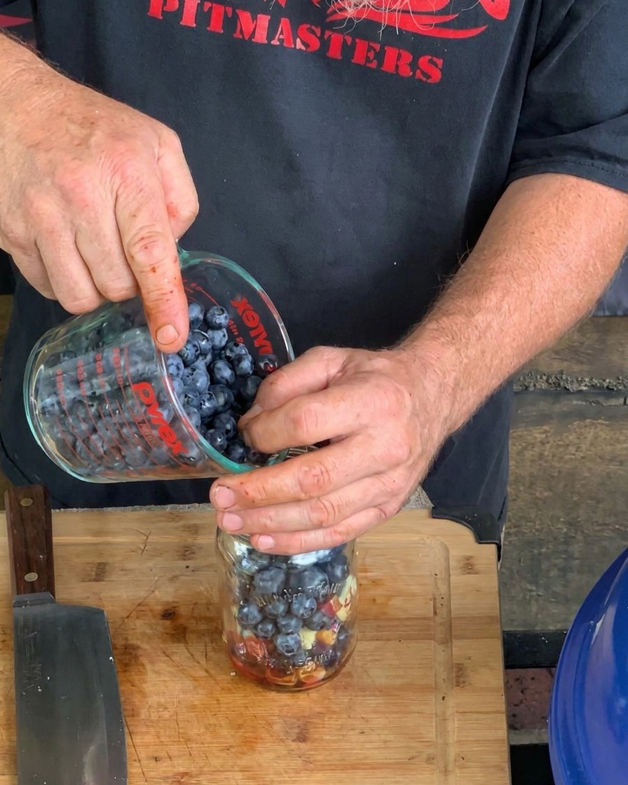 Primo piano di mani che versano mirtilli frescos da un misuratore di vetro in un barattolo di vetro che contiene altri ingredienti liquidi e solidi per la salsa.