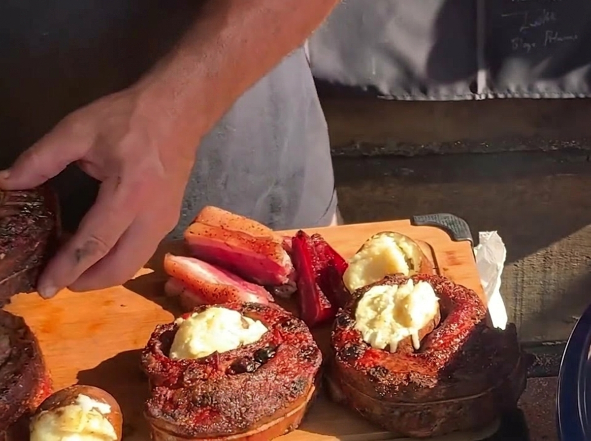 Una mano inserisce una patata arrosto calda e imburrata nella cavità centrale di un cestino di costine cotto e rotondo su un tagliere di legno.