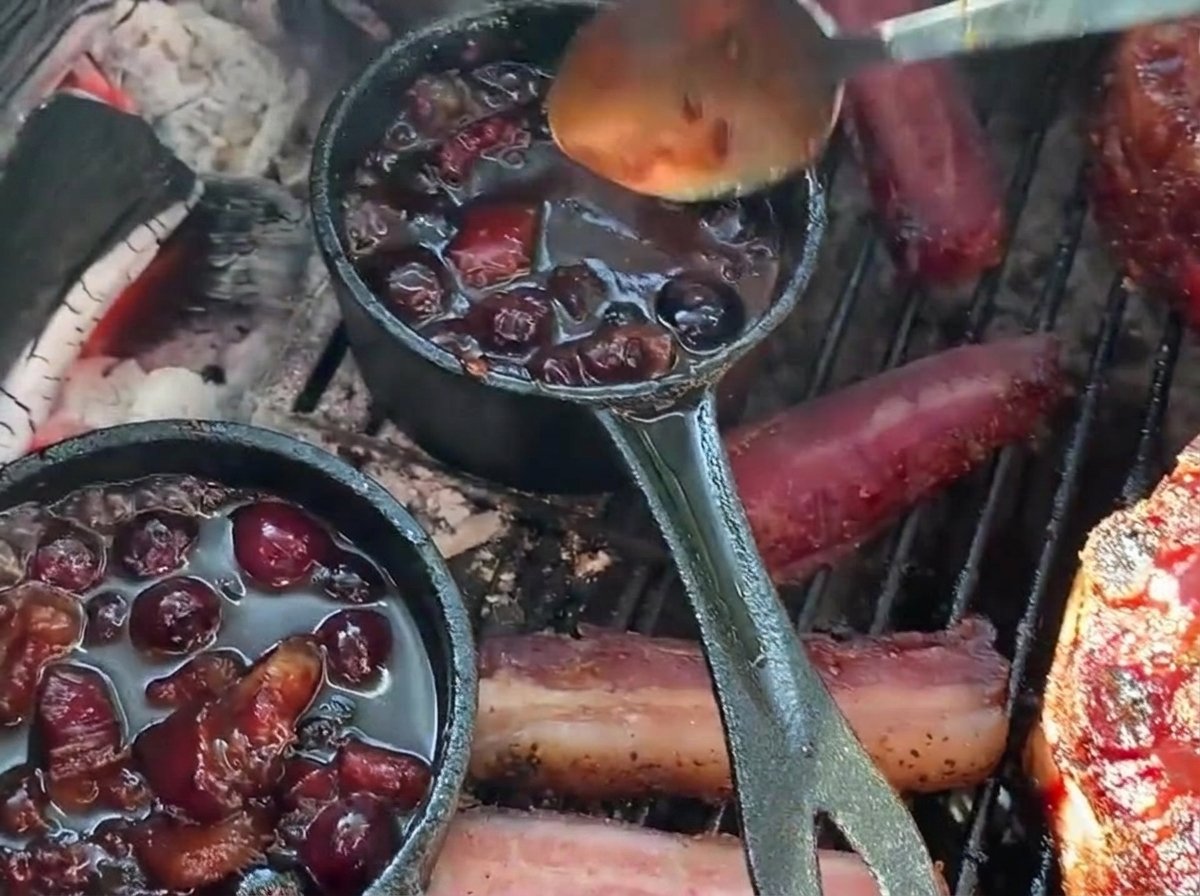 Un cucchiaio versa una ricca salsa barbecue ai mirtilli da un pentolino in ghisa su un cestino di costine farcito sul barbecue.