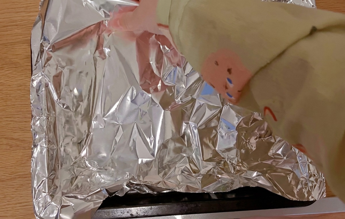 Mani che coprono le costine di maiale marinate su una teglia da forno nera con un foglio di alluminio lucido.