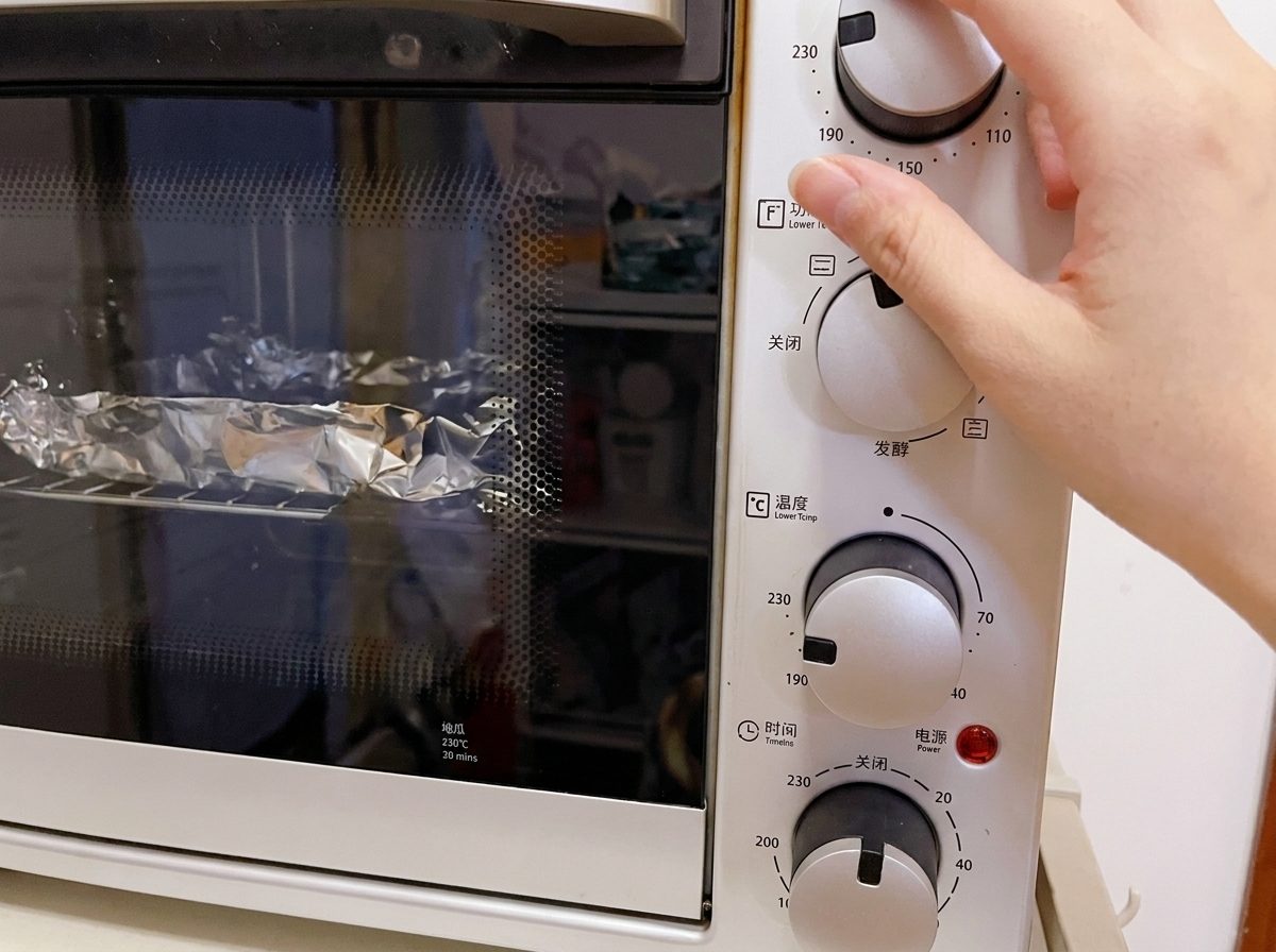 Primo piano di una mano che regola la manopola della temperatura di un fornetto elettrico bianco a 230°C, con i cartocci in alluminio visibili sulla griglia interna.