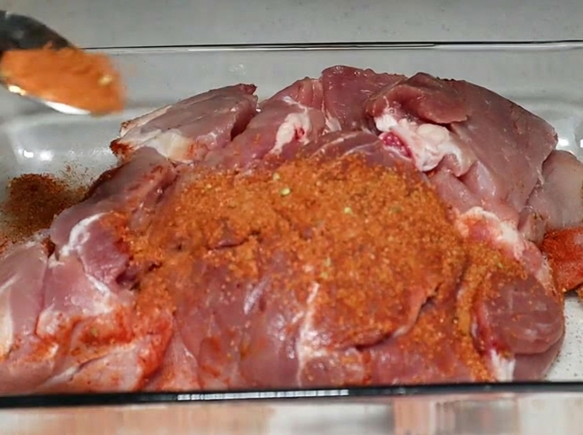 Un cucchiaio colmo di spezie rossastre viene cosparso su un grosso pezzo di spalla di maiale cruda adagiata in una pirofila di vetro.