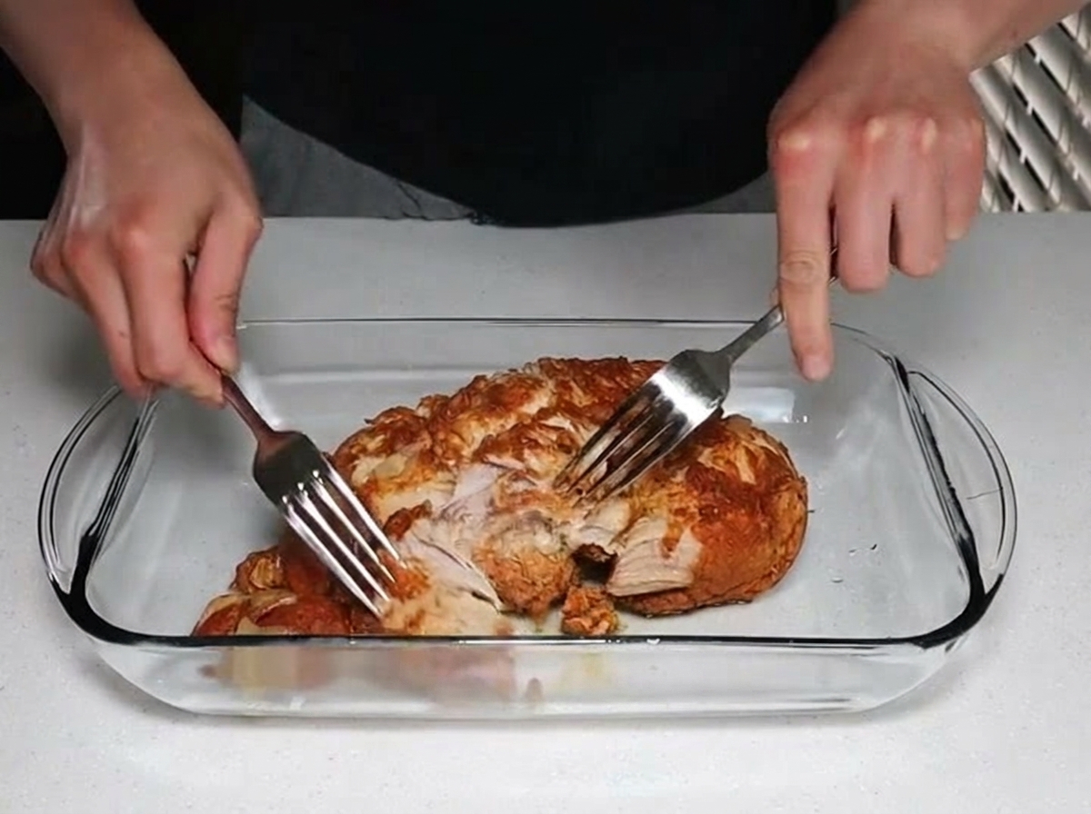 Due mani utilizzano delle forchette in acciaio inox per tirare e sfilacciare un abbondante pezzo di morbido maiale cotto allinterno di una pirofila in vetro.