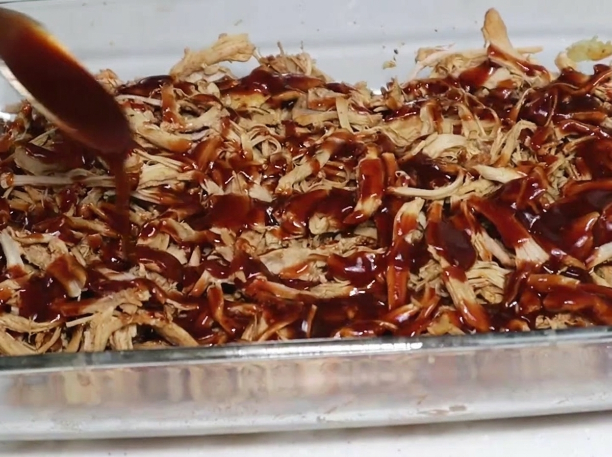 Una cucchiaiata di salsa barbecue densa e scura cola sopra uno strato di maiale sfilacciato affumicato in una pirofila di vetro trasparente.