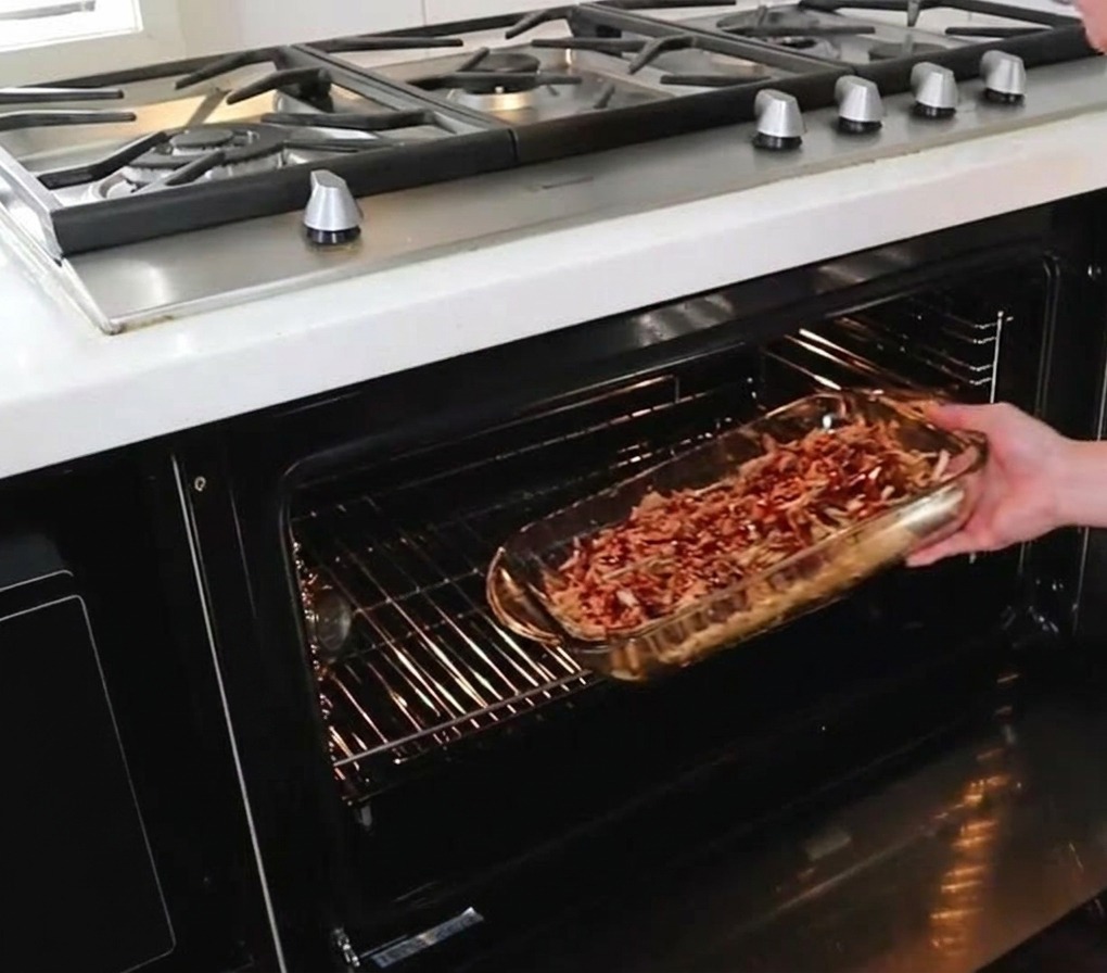 Una persona inforna una pirofila di vetro ricolma di pulled pork condito con salsa, facendola scivolare sulla griglia centrale del forno caldo.