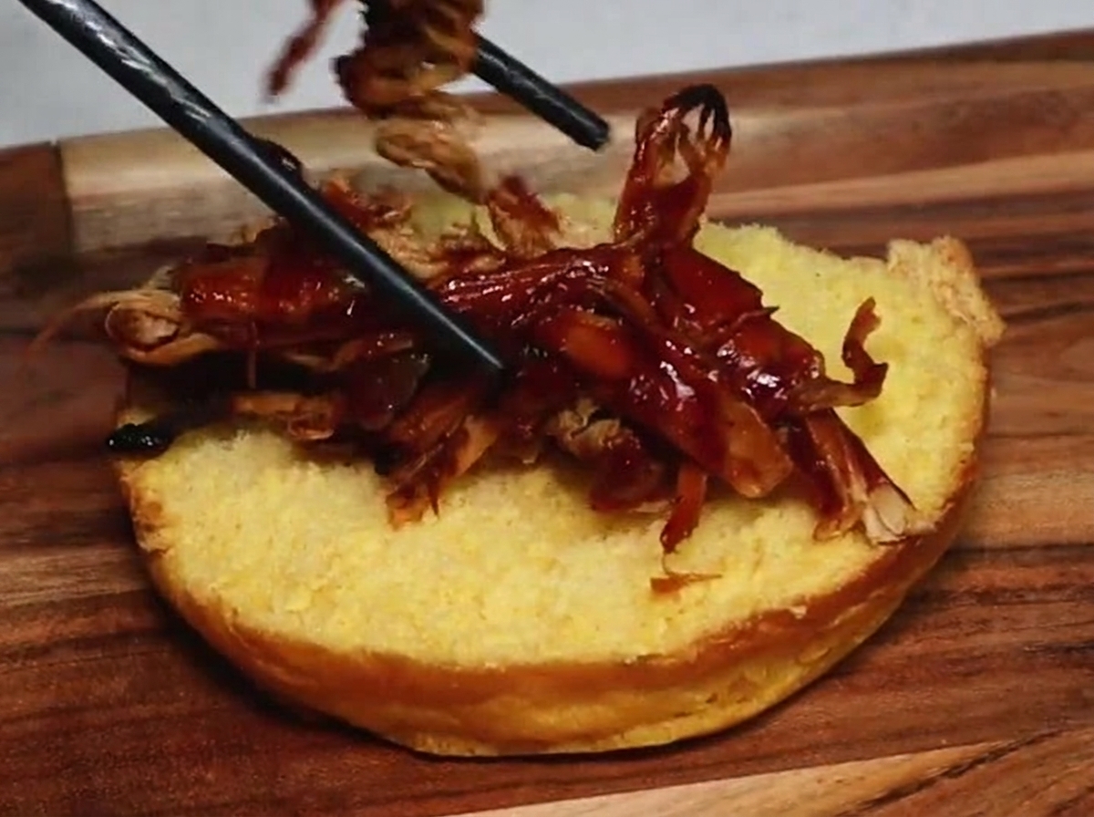 Delle bacchette appoggiano succulenti sfilacci di pulled pork, avvolti da una salsa barbecue scura, sulla superficie dorata di metà panino tostato.