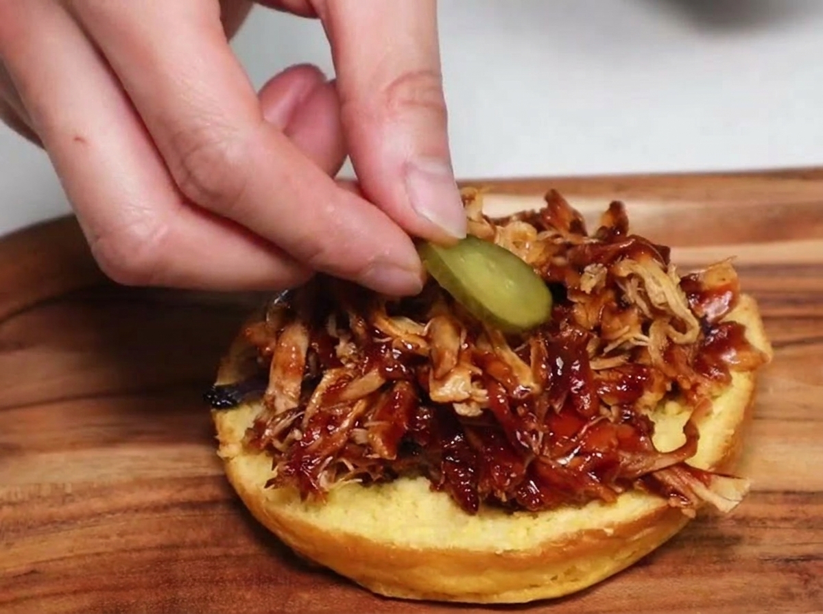 Una mano appoggia una fetta rotonda di cetriolino sottaceto sopra una montagnola di maiale sfilacciato al barbecue, su un panino tostato.