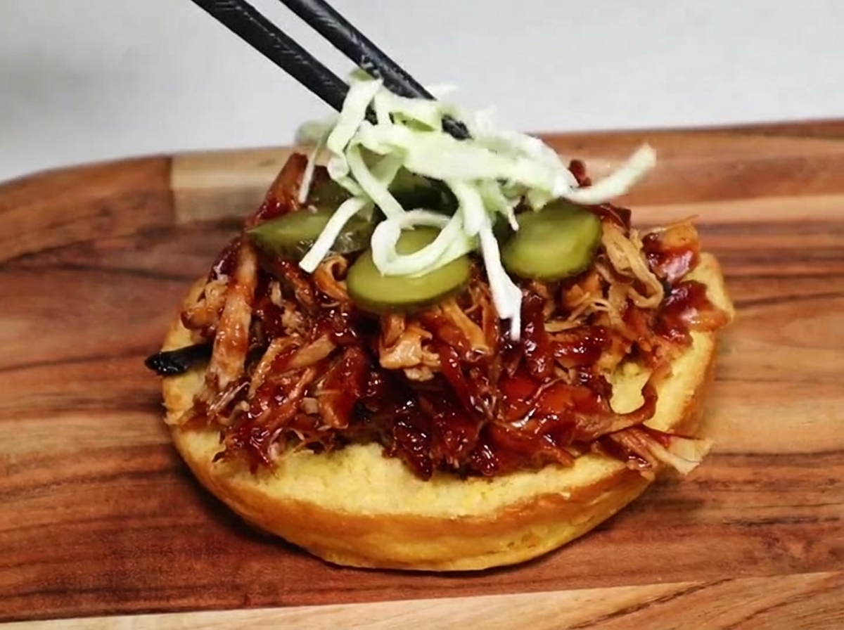 Bacchette posizionano dellinsalata coleslaw di cavolo verde sopra le fette di cetriolino e il pulled pork in un panino da hamburger.