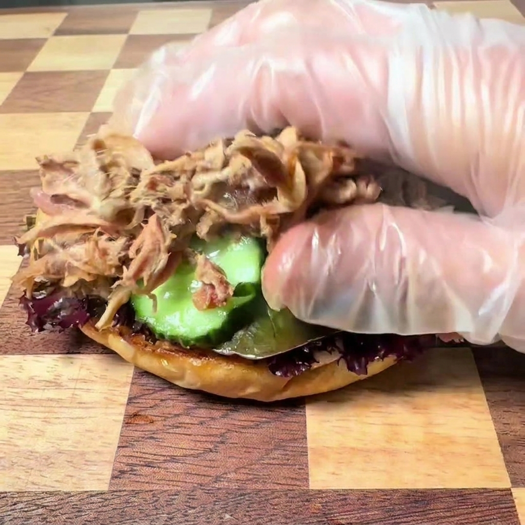 Una mano con un guanto di plastica trasparente che adagia del tenero pulled pork sfilacciato sulla base del burger con lattuga e fette di cetriolo.
