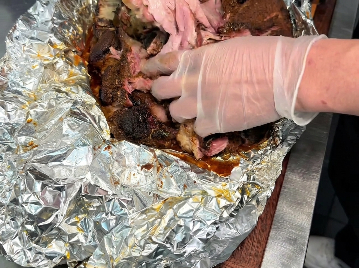 Mani con guanti che sfilacciano un grosso pezzo di tenero maiale affumicato cotto su una superficie rivestita di alluminio.