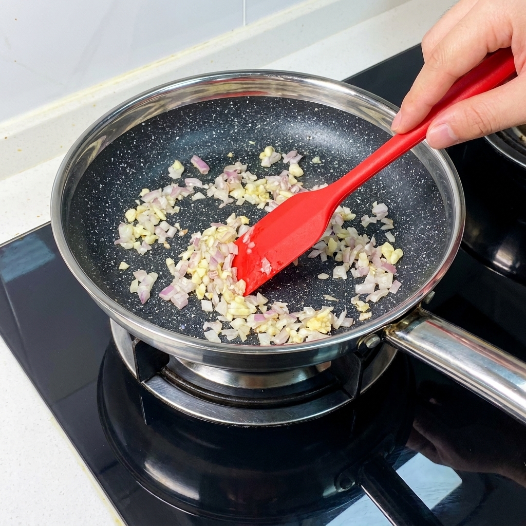 Cipolle rosse sminuzzate finemente e aglio tritato che appassiscono in una padella, mescolati con una spatola rossa.