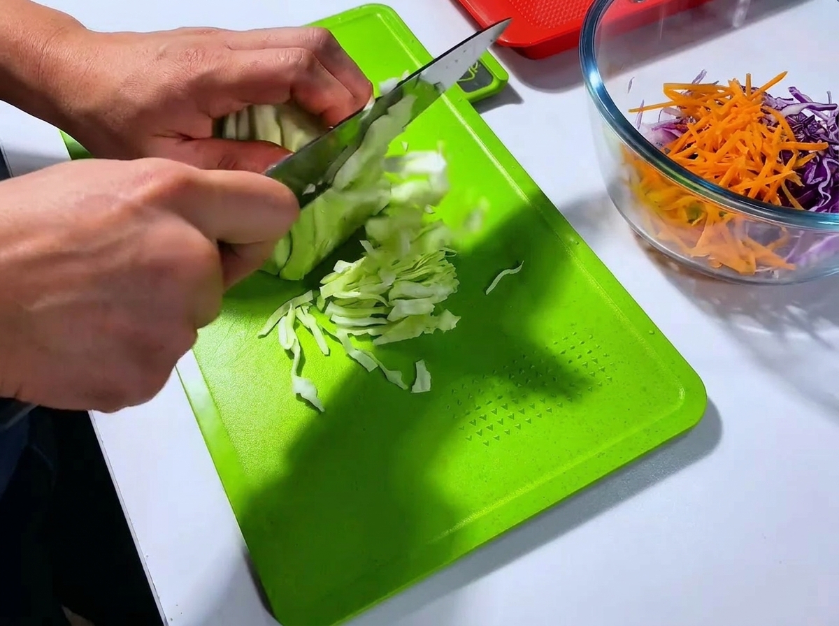 Un cuoco che affetta finemente un cespo di cavolo cappuccio verde con un grande coltello su un tagliere verde brillante.